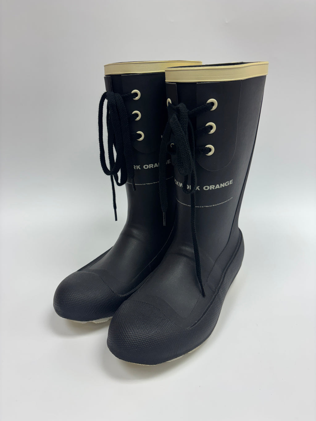 A pair of black rubber rain boots with black laces, gold eyelets, and a beige inner lining, shown on a light gray background.