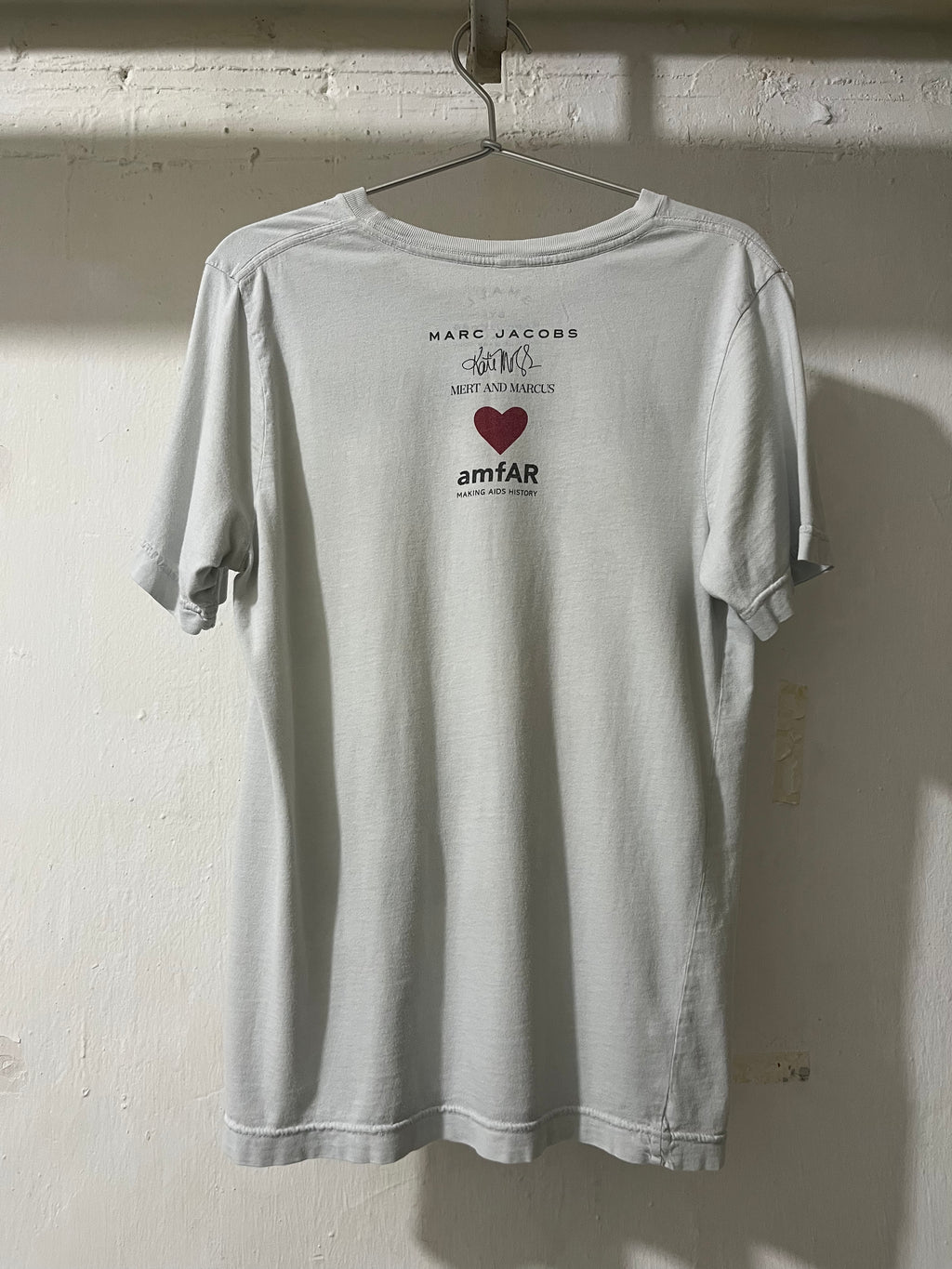 Gray t-shirt with Marc Jacobs and amfAR logos on a hanger against a light-colored wall.