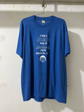A blue short-sleeve graphic t-shirt hanging on a metal hanger against a white textured wall. The shirt features a white printed text graphic on the front reading 'I had a MAD time at Matthe & party March 24, 19' above a small white crest graphic.