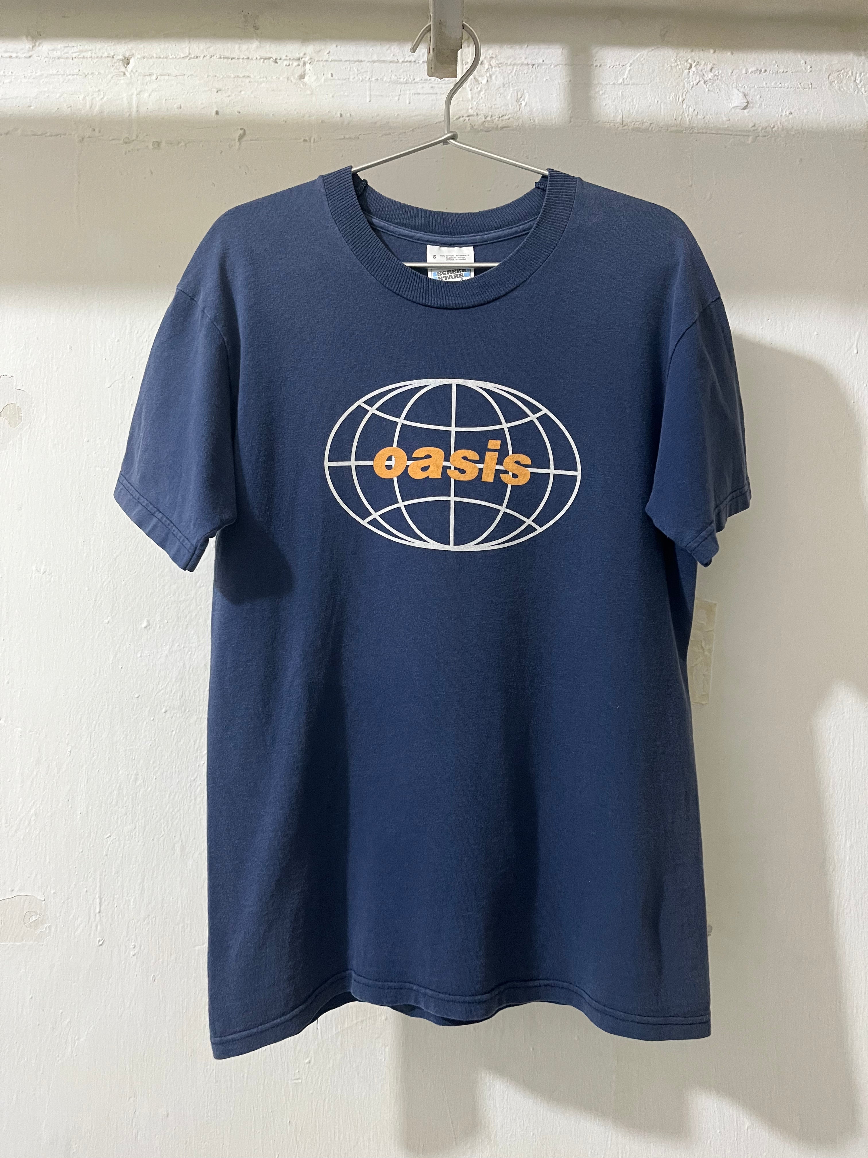 A navy blue short-sleeve t-shirt hanging on a metal hanger against a white brick wall. The shirt features a white globe graphic on the chest with the word 'oasis' printed in orange across the center.