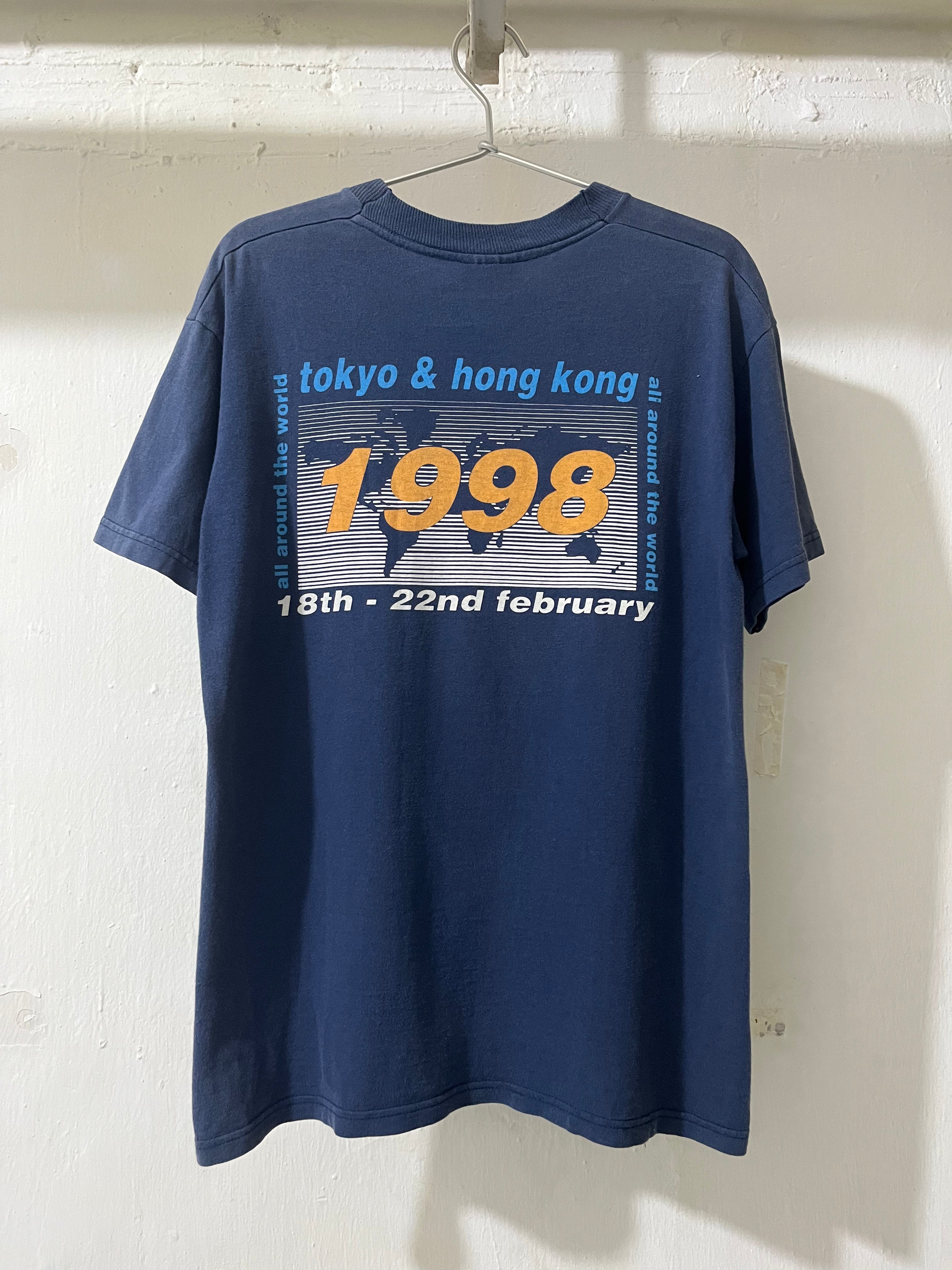 A navy blue short-sleeve t-shirt hanging on a metal hanger against a white wall. The back of the shirt features a graphic print with the text 'tokyo & hong kong', a large yellow '1998', and '18th - 22nd february' over a gray and white striped map design.