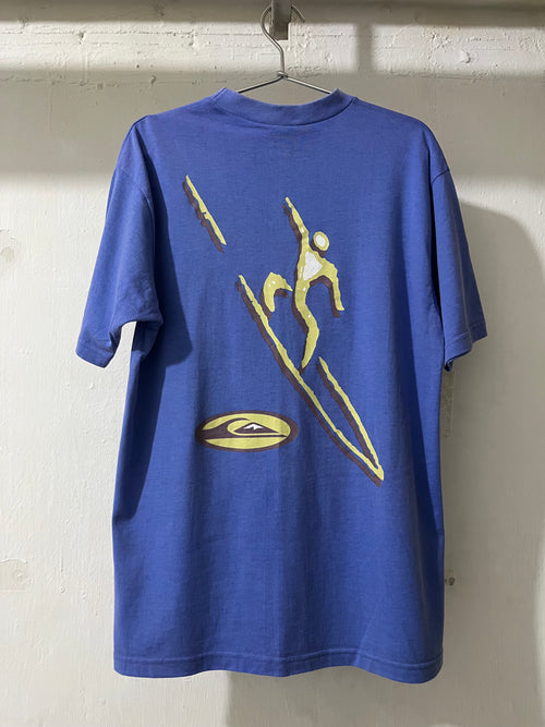 A blue short-sleeve t-shirt hanging on a metal hanger against a white brick wall, featuring a large yellow graphic print of a surfer riding a board on the back.