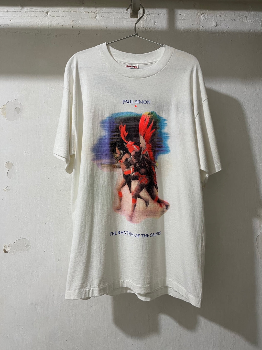 An oversized off-white graphic t-shirt hanging on a metal hanger against a textured white wall. The shirt features a colorful print of a winged figure with red hair and blue text reading 'INNI SIMON' and 'THE RHYTHM OF THE ANXIETY'.