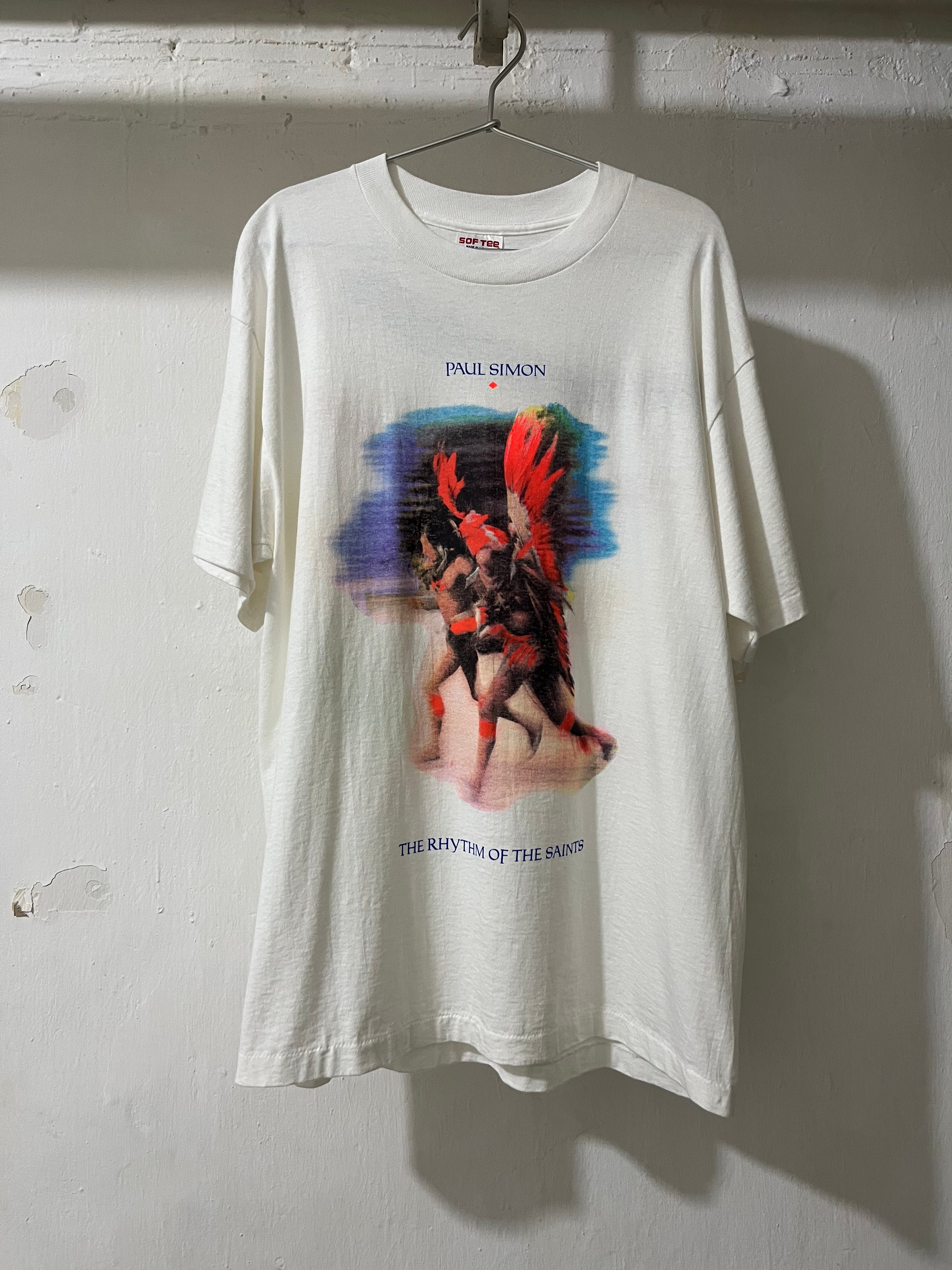 An oversized off-white graphic t-shirt hanging on a metal hanger against a textured white wall. The shirt features a colorful print of a winged figure with red hair and blue text reading 'INNI SIMON' and 'THE RHYTHM OF THE ANXIETY'.