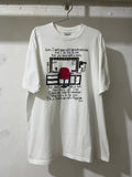 A white graphic t-shirt hanging on a metal hanger against a white painted brick wall. The t-shirt features a black and red cartoon illustration of a computer desk setup with text above and below the graphic.