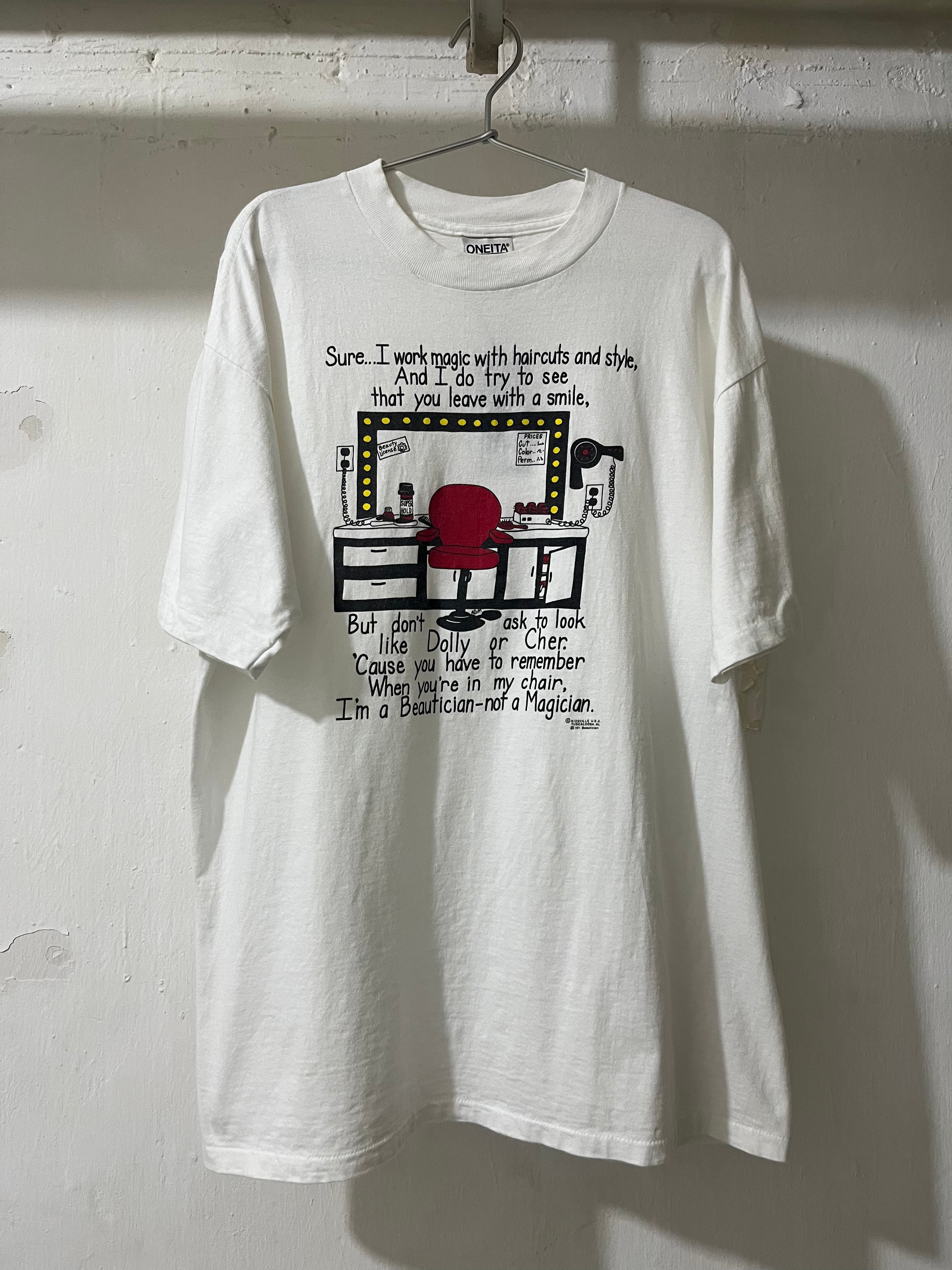 A white graphic t-shirt hanging on a metal hanger against a white painted brick wall. The t-shirt features a black and red cartoon illustration of a computer desk setup with text above and below the graphic.