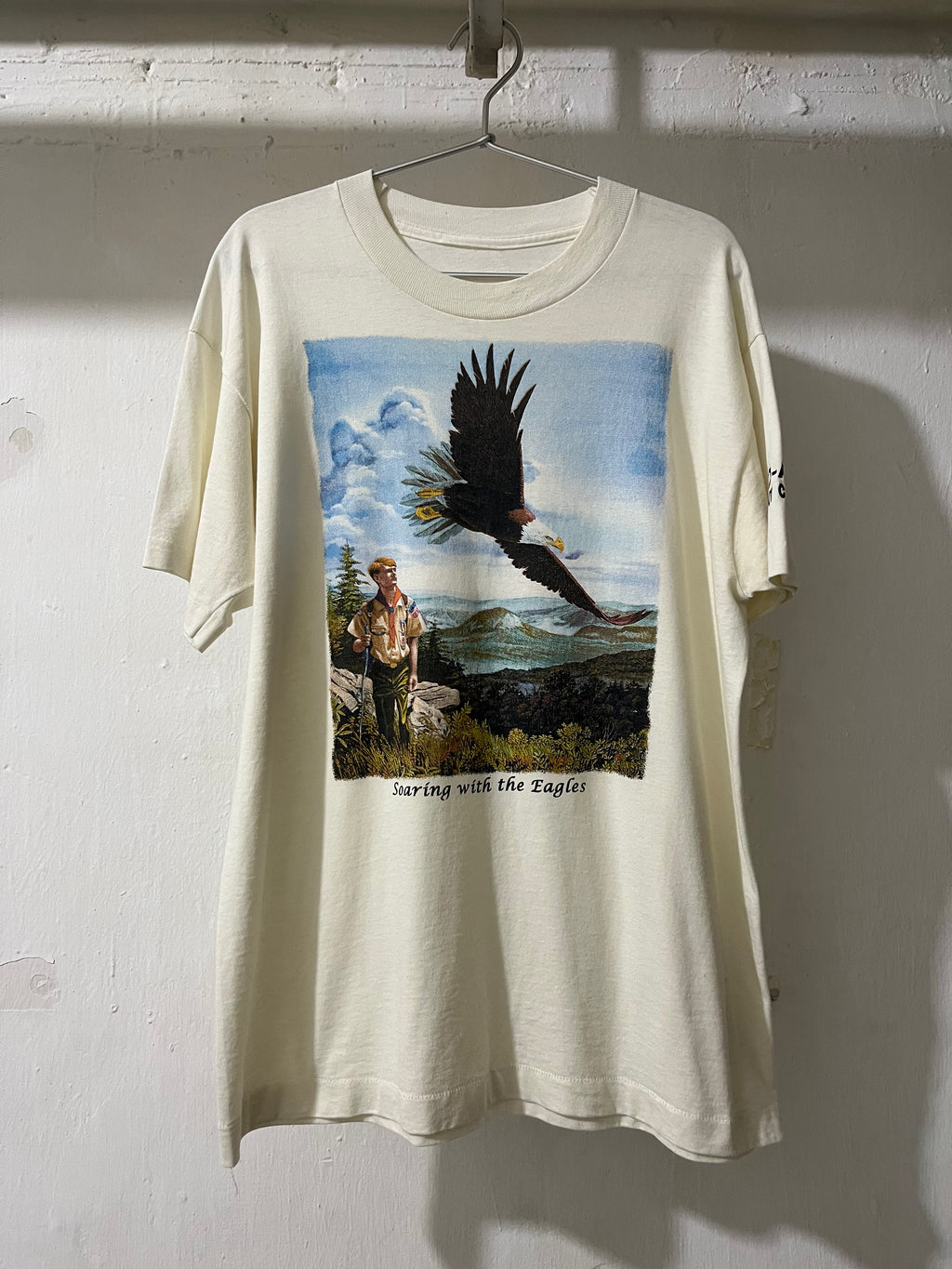 An off-white short-sleeve t-shirt hanging on a metal hanger against a white wall. The shirt features a large graphic print on the front depicting a bald eagle flying over a man standing on a rocky cliffside, with the text 'Surfing with the Eagles' printed below the scene.