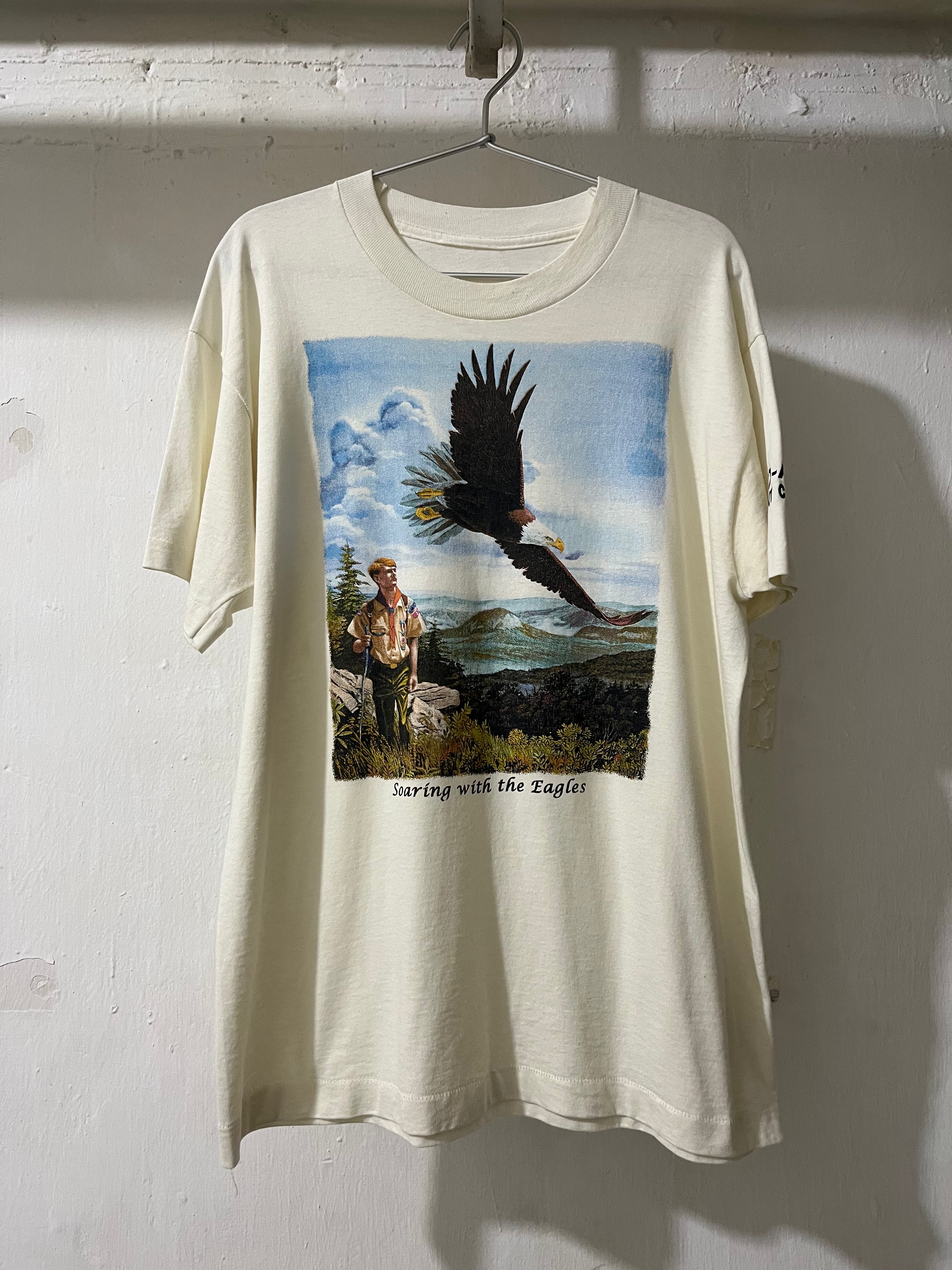 An off-white short-sleeve t-shirt hanging on a metal hanger against a white wall. The shirt features a large graphic print on the front depicting a bald eagle flying over a man standing on a rocky cliffside, with the text 'Surfing with the Eagles' printed below the scene.