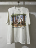 A white graphic t-shirt hanging on a metal hanger against a textured white wall. The shirt features a printed illustration of two deer under trees with the text 'THE DEER' and 'Bummer of a birthmark Hol!'.