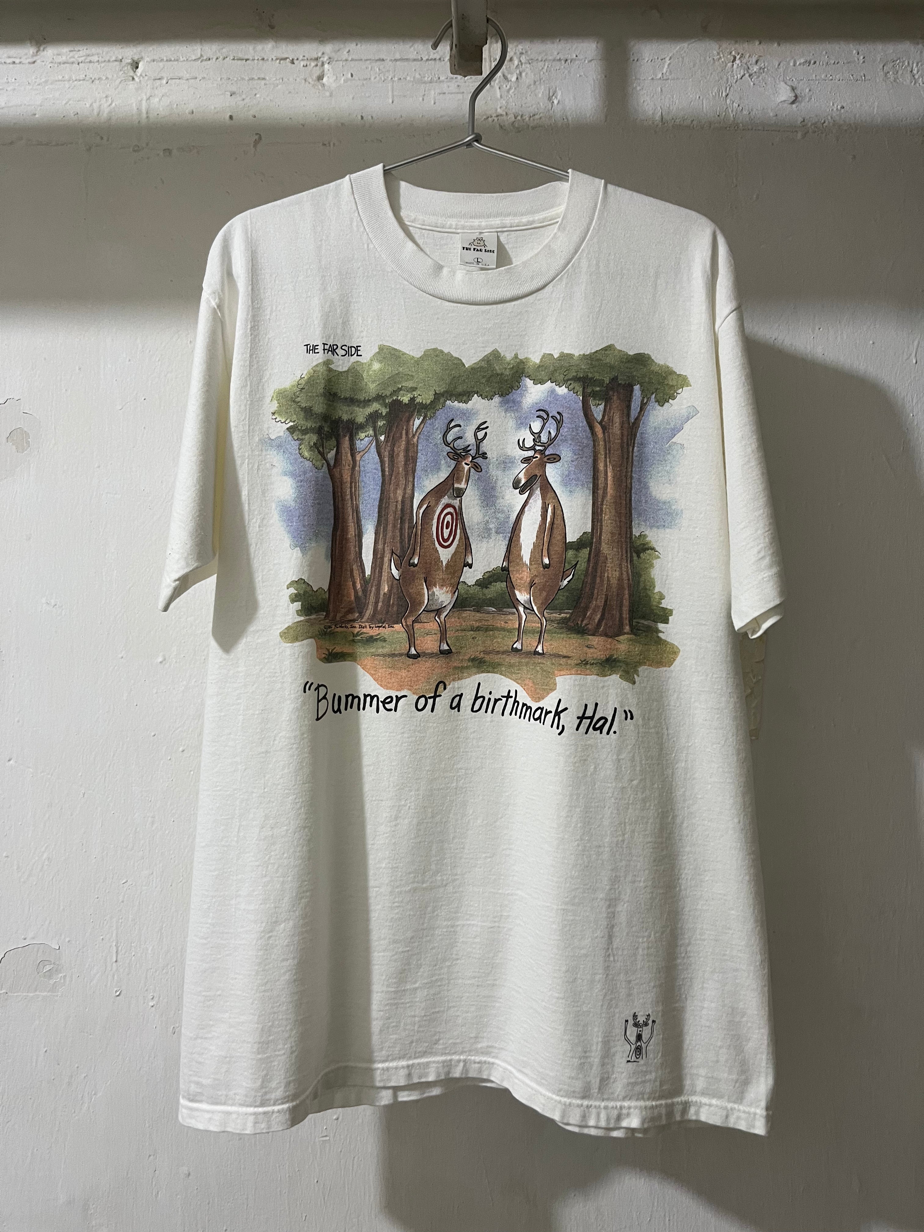 A white graphic t-shirt hanging on a metal hanger against a textured white wall. The shirt features a printed illustration of two deer under trees with the text 'THE DEER' and 'Bummer of a birthmark Hol!'.