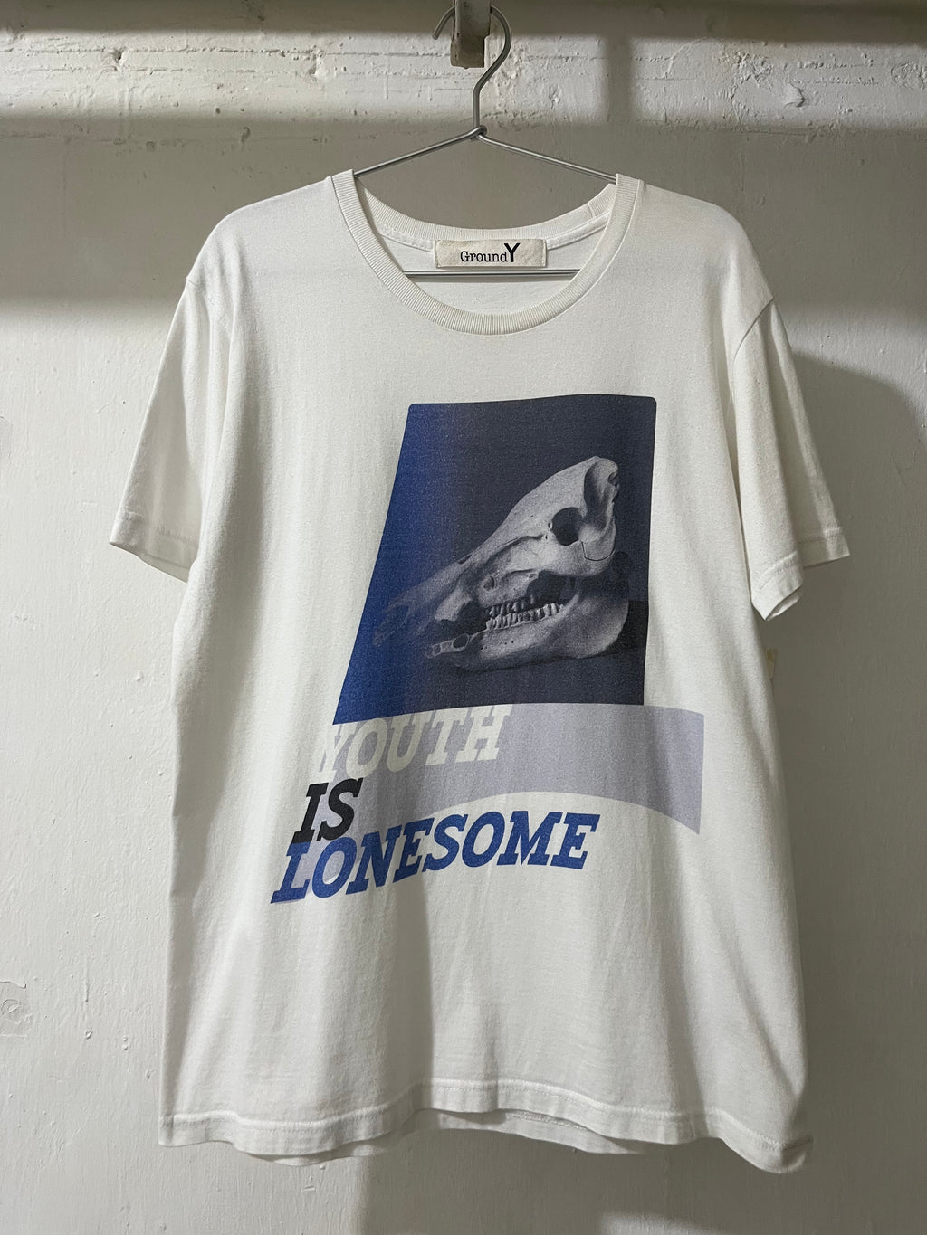 An off-white short-sleeve t-shirt hanging on a metal hanger against a white brick wall. The shirt features a graphic print of a dog skull inside a dark blue square, with the text 'YOUTH IS LONESOME' printed below in blue and gray.