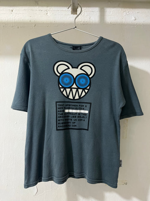 A faded slate blue t-shirt hanging on a metal hanger against a white painted brick wall. The shirt features a graphic of a cartoon bear face with blue eyes and a sharp toothy grin, above a black text box graphic.