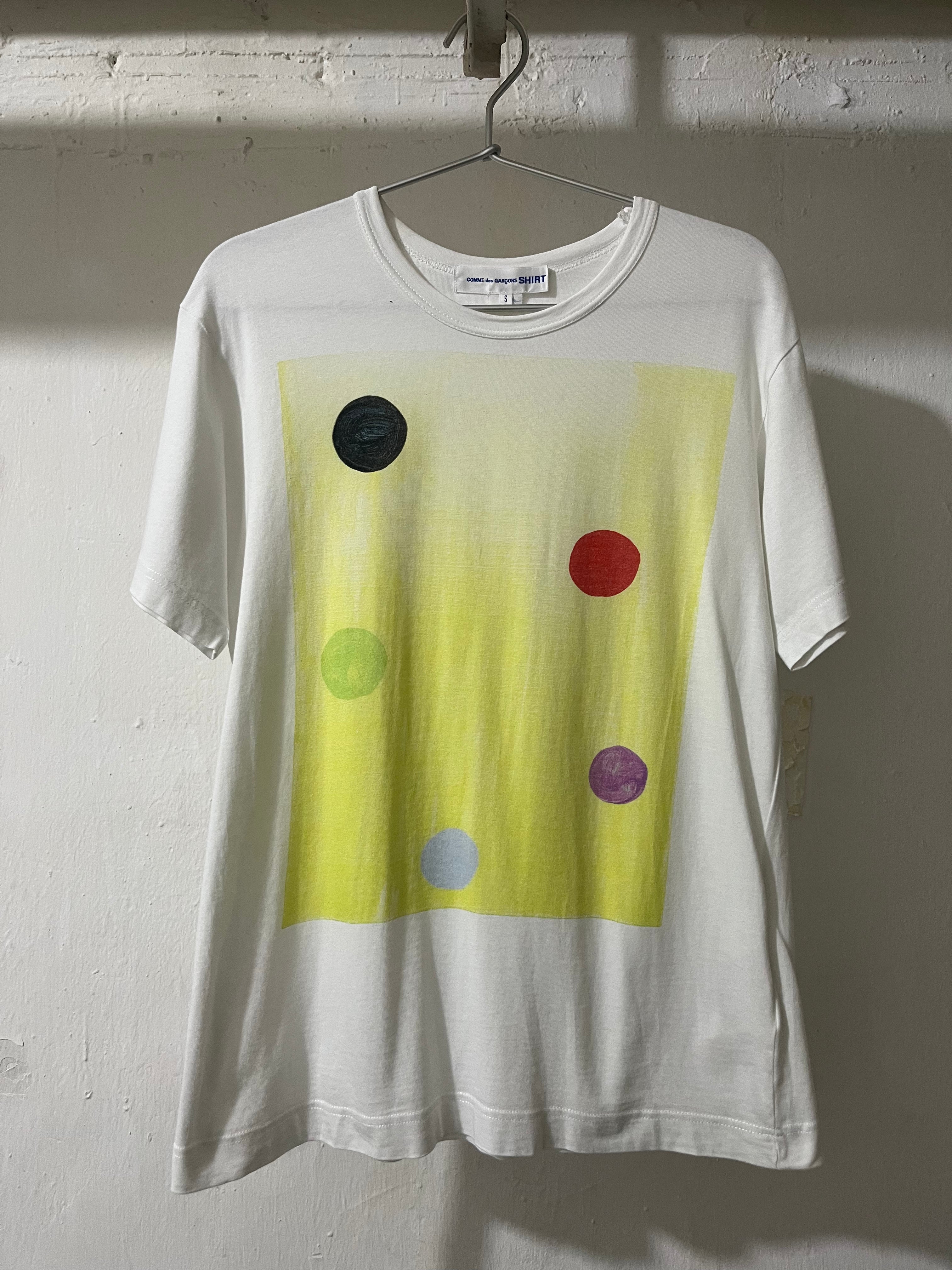 A white short-sleeve t-shirt hanging on a metal hanger against a textured white wall. The shirt features a large yellow rectangular graphic on the front with several colorful painted dots in black, green, red, purple, and light blue.