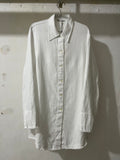 A white long-sleeve button-up shirt with a pointed collar, hanging on a metal hanger against a textured, off-white wall.