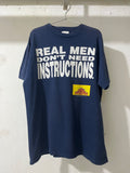 A navy blue oversized t-shirt hanging on a metal hanger against a white wall. The shirt features the text 'REAL MEN DON'T NEED INSTRUCTIONS.