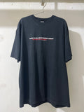 A faded black oversized short-sleeve t-shirt hanging on a metal hanger against a white textured wall. The shirt features a small graphic text print on the chest in white and red.