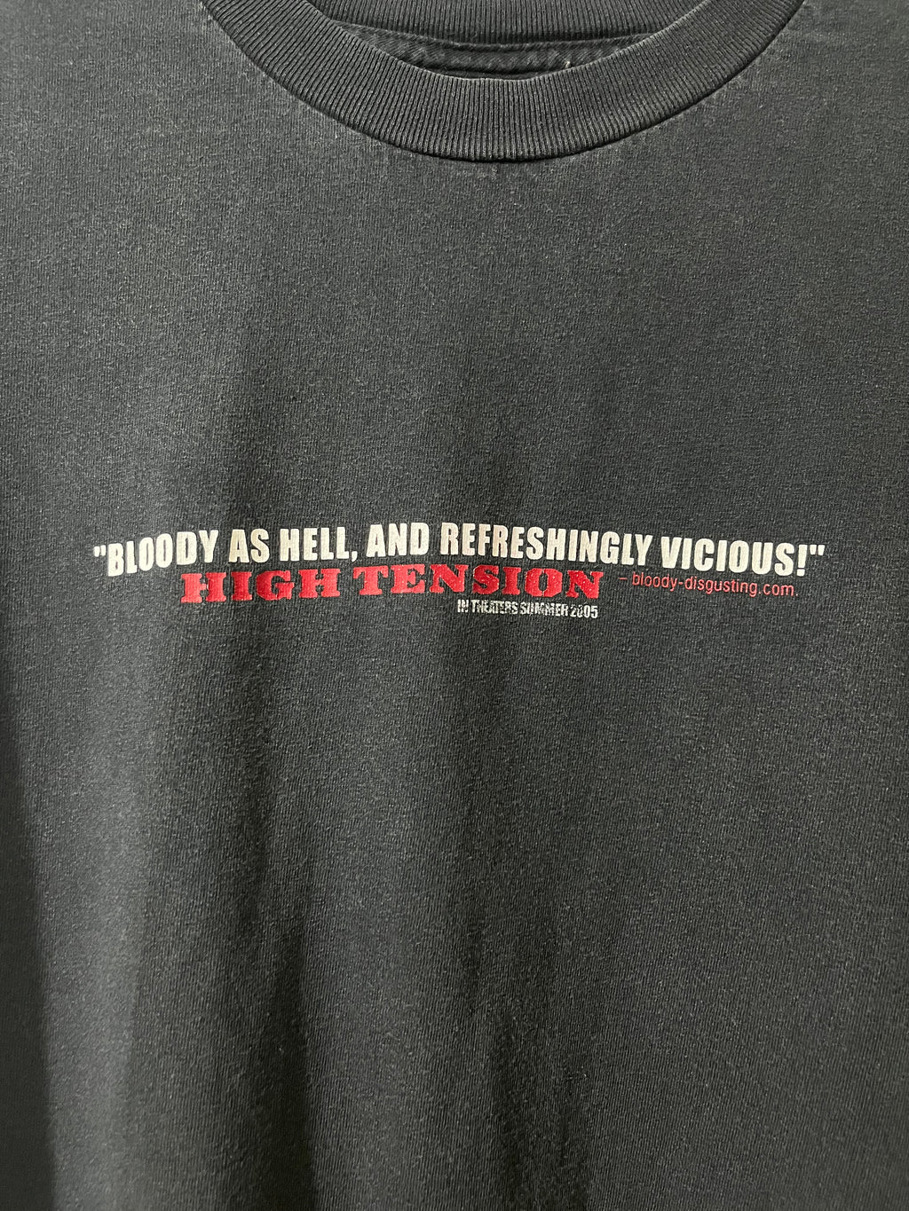 Close-up of a dark charcoal gray t-shirt featuring a graphic print on the chest with the text 'BLOODY AS HELL, AND REFRESHINGLY VICIOUS!' in white, 'HIGH TENSION' in red, and 'Bloody Shipping.com' in red and white.