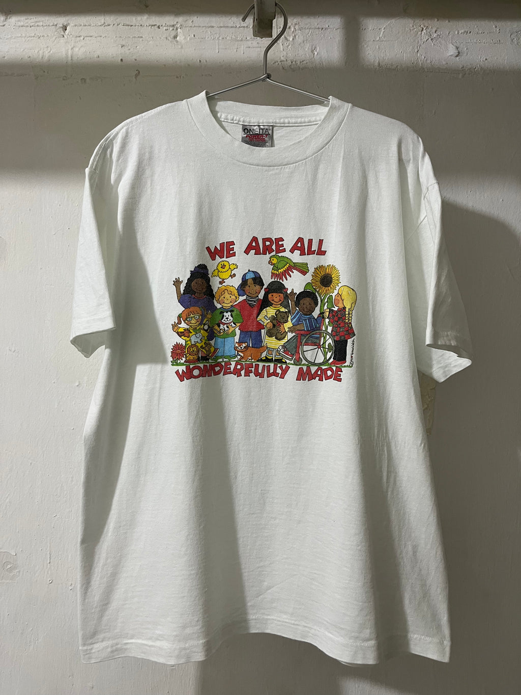 A white short-sleeve graphic t-shirt hanging on a metal hanger against a white painted brick wall. The front of the shirt features a colorful cartoon illustration of diverse children and the text 'WE ARE ALL WONDERFULLY MADE' in red.