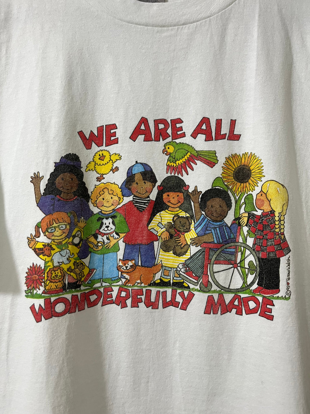 A white short-sleeve t-shirt featuring a colorful graphic of diverse children, pets, and a wheelchair with the text 'WE ARE ALL WONDERFULLY MADE' in red.