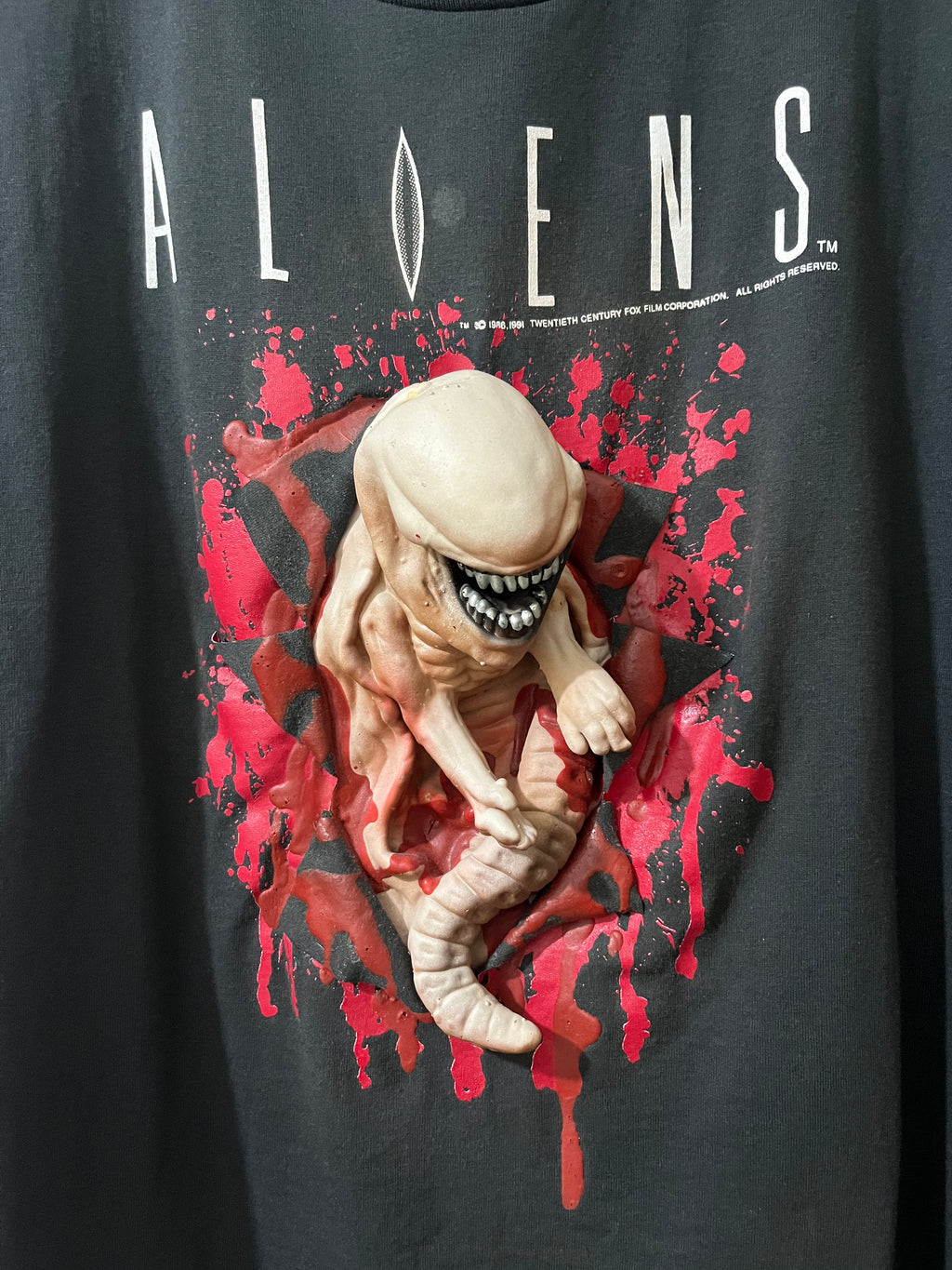 A close-up of a black graphic t-shirt featuring a 3D-printed Alien figure bursting out of a red blood-splatter design, with the word 'ALIENS' printed in white across the chest.
