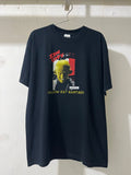 A black graphic t-shirt hanging on a metal hanger against a white brick wall. The shirt features a yellow and red graphic print of a bald man's face with the text 'SLAY THE BASTARD' in red and yellow.