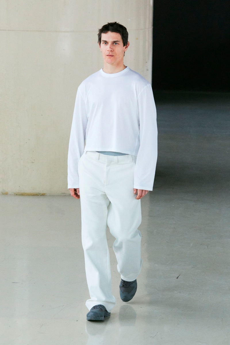 A male model walks down a runway wearing a white long-sleeve cropped t-shirt, white trousers, and dark gray slip-on shoes.