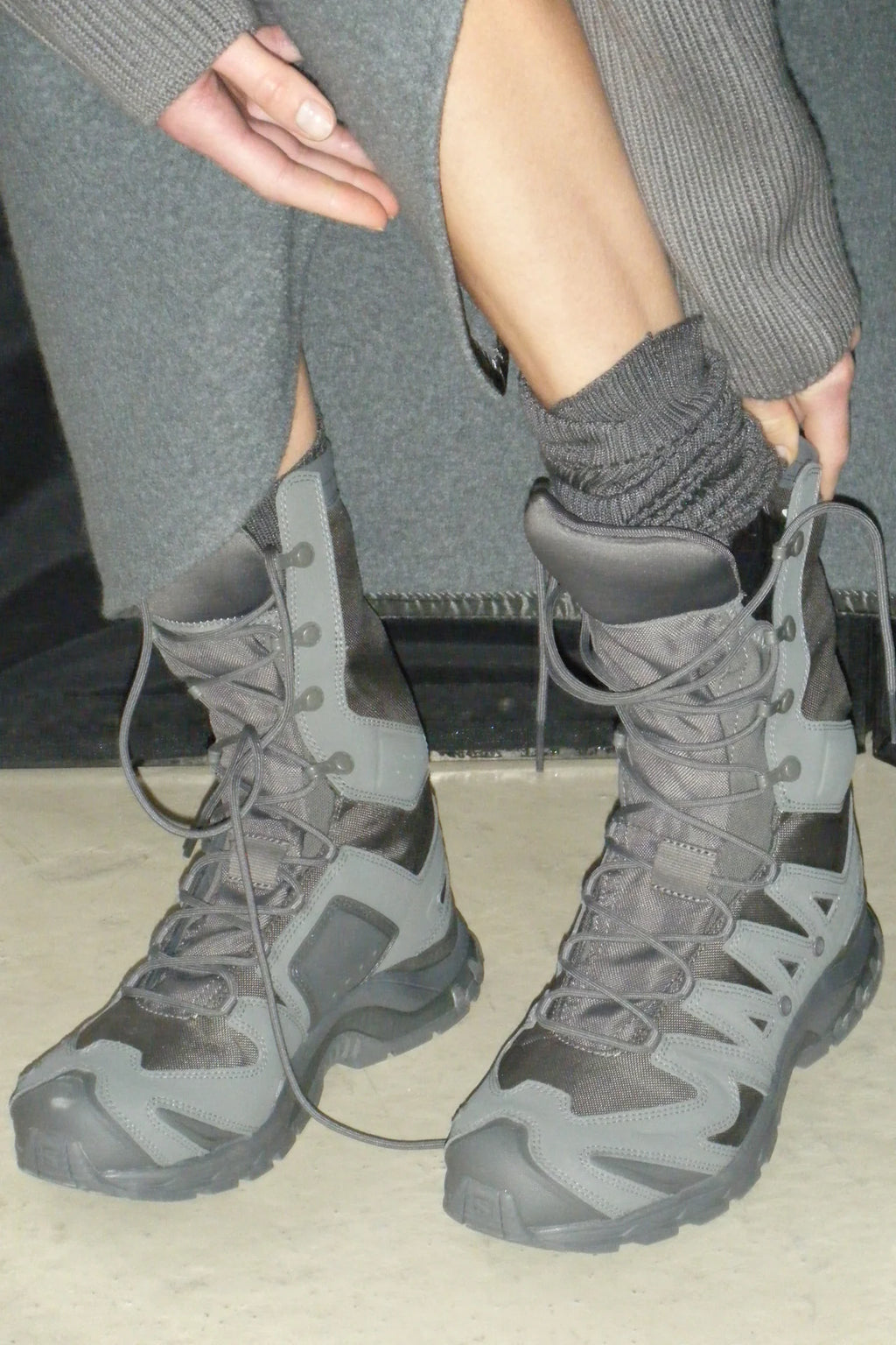A person wearing gray tactical-style lace-up boots with gray knit socks, seated indoors near a dark bench or chair.