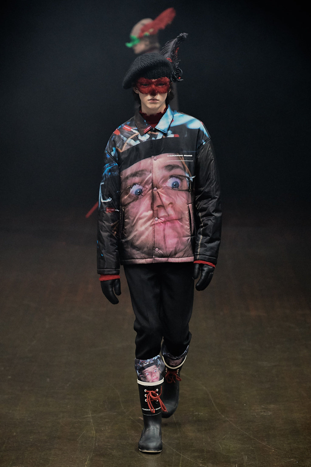 A model walks the runway wearing a dark jacket with a large surreal face print on the front, black pants, black boots, and a black hat with red and green accents.