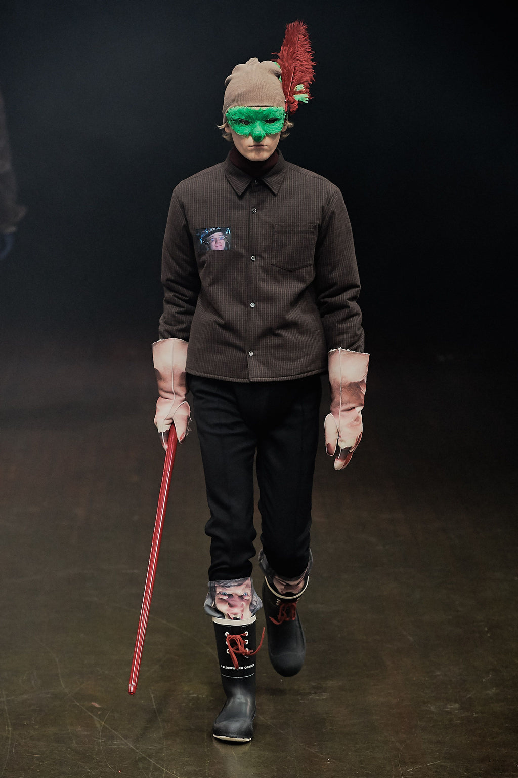 A model walks the runway wearing a dark brown button-up shirt, black trousers, and black rain boots with pink and red patterned socks. The outfit is accessorized with a beige knit beanie featuring a red feather, green-tinted face masks, and pink gloves, while holding a red cane against a dark runway background.